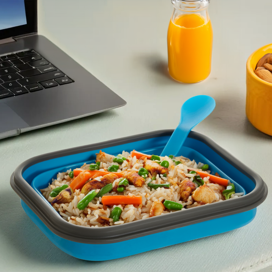 Blue and gray food container with rice and vegetables on a desk with a laptop and juice bottle.