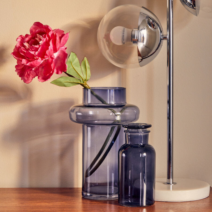Glass vase with a pink flower on a wooden surface next to a lamp.