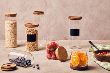 Set of glass jars with wooden lids on a table with fruits and a bowl.