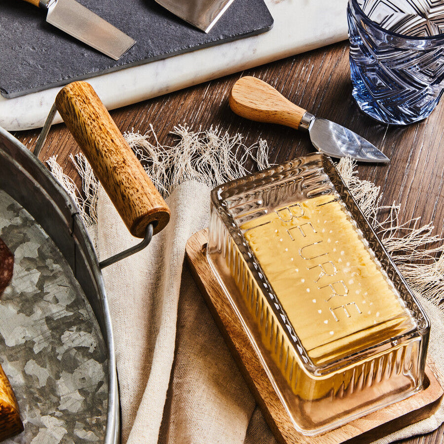 Butter dish with wooden spreader, cheese knives, and a glass container on a wooden surface.