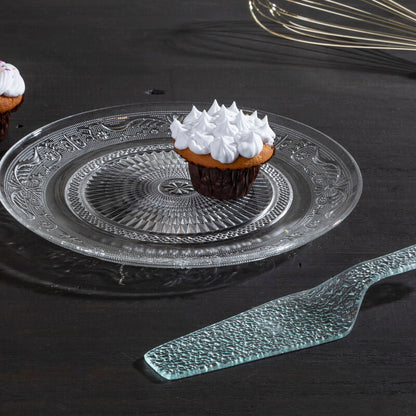 Decorative glass plate with a cupcake on a dark surface, accompanied by a whisk and knife.