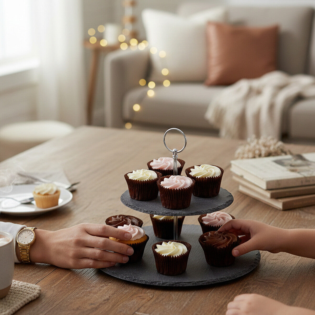 Two people reaching for cupcakes on a tiered stand in a cozy living room.