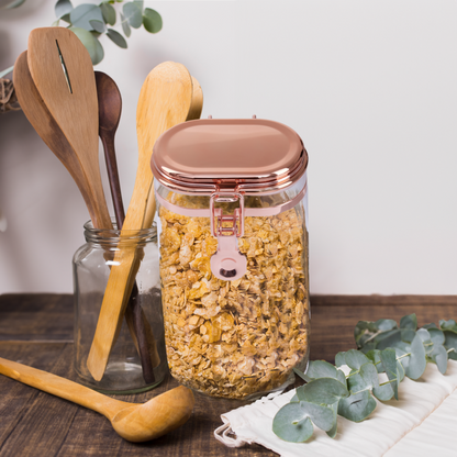 Clear jar with rose gold lid containing granola on a wooden surface with wooden spoons and eucalyptus leaves.