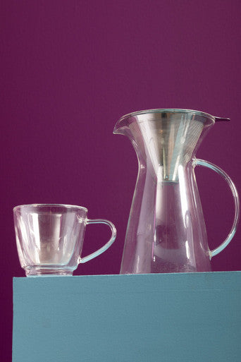 Clear glass pitcher and cup on a teal surface with a purple background