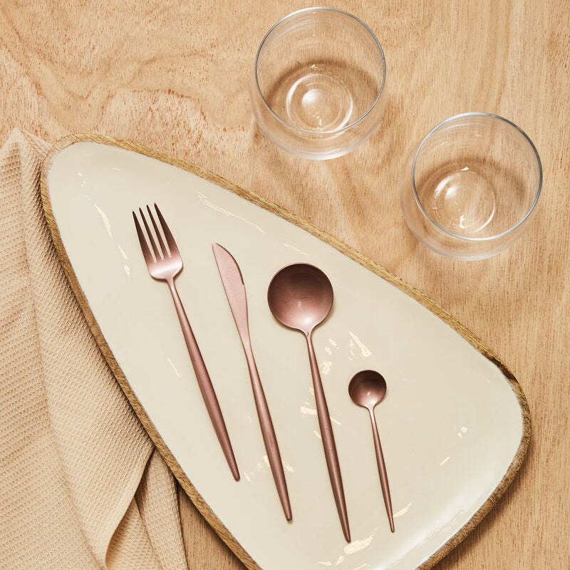 Rose gold cutlery set on a ceramic plate with two clear glasses on a wooden surface.