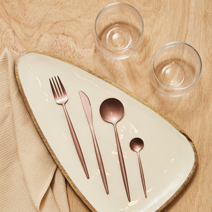 Rose gold cutlery set on a ceramic plate with two clear glasses on a wooden surface.