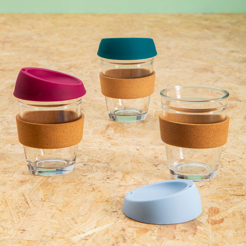 Set of reusable glass cups with silicone sleeves and lids on a wooden surface.