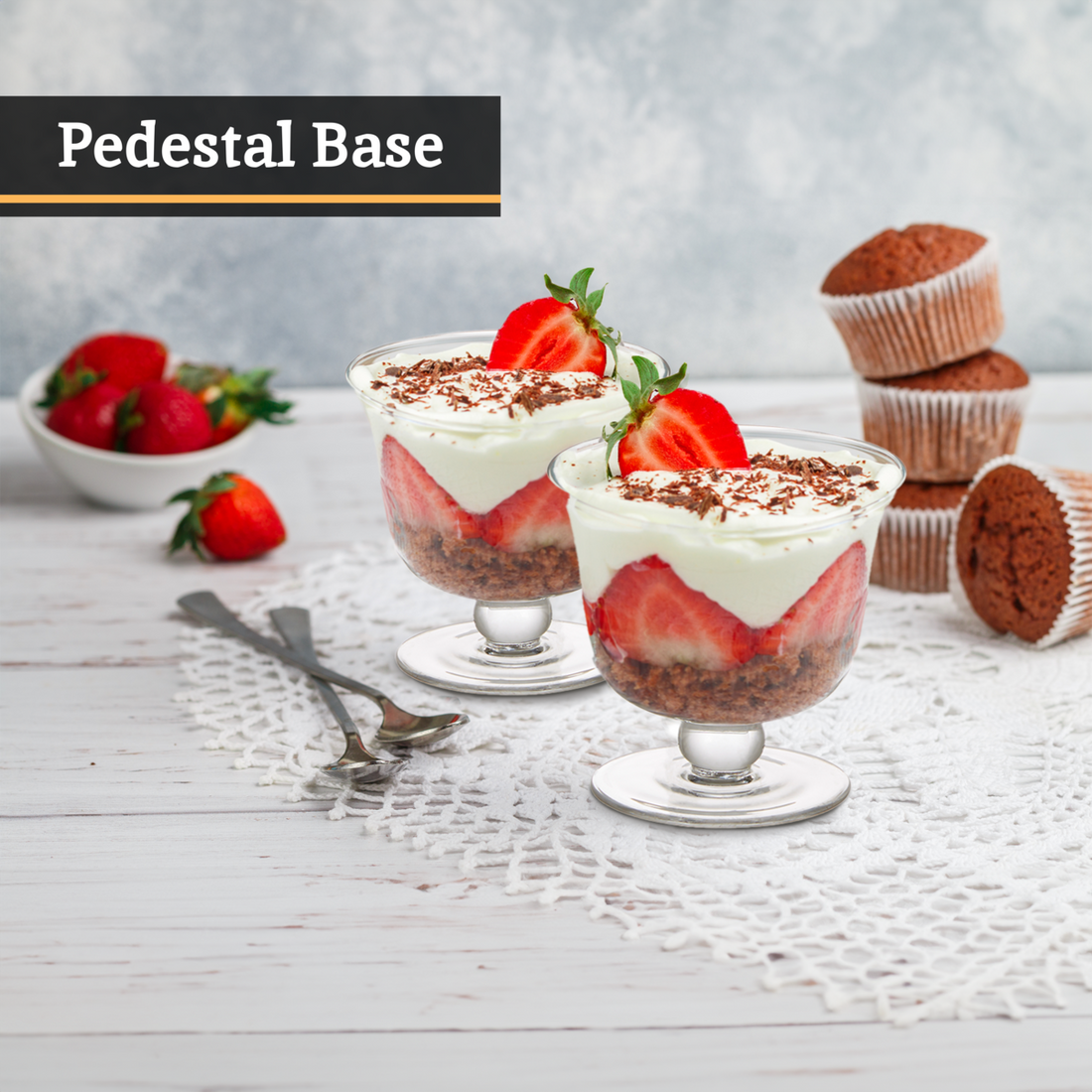 Two dessert glasses with strawberries and whipped cream on a wooden table with cupcakes and strawberries.