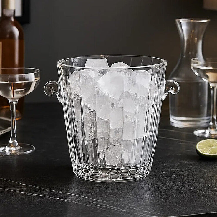 Glass ice bucket with ice cubes on a bar counter with cocktail ingredients.