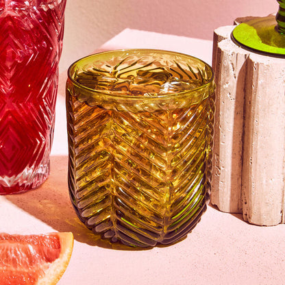 Two textured glass tumblers with red and green drinks, accompanied by a slice of grapefruit on a pink surface.