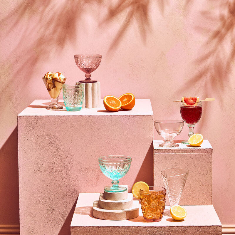 Cocktail setup with glasses and fruits on a pink surface with palm leaf shadows.