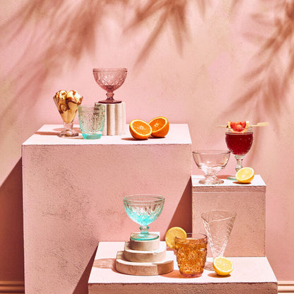 Cocktail setup with glasses and fruits on a pink surface with palm leaf shadows.