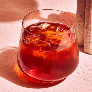 Glass of red cocktail with ice and orange slices on a light pink surface