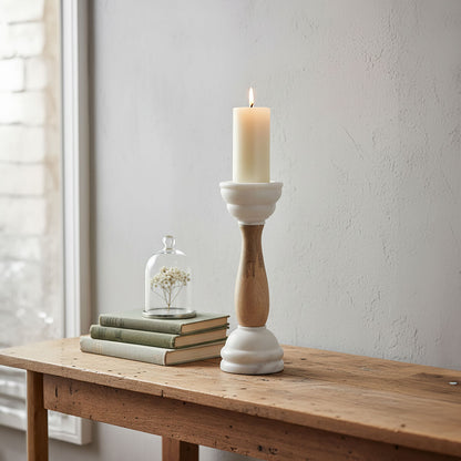 Candle in a wooden and white candlestick on a wooden table with books and a small plant dome in the background.
