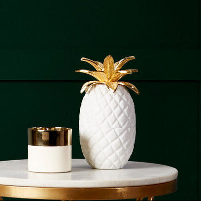Small round table with gold legs and marble top, featuring a decorative pineapple and candle against a dark green wall.