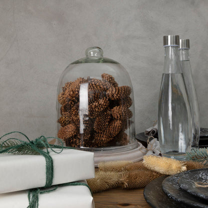 Decorative setting with pinecones under a glass dome, wrapped gifts, and a bottle on a wooden surface.