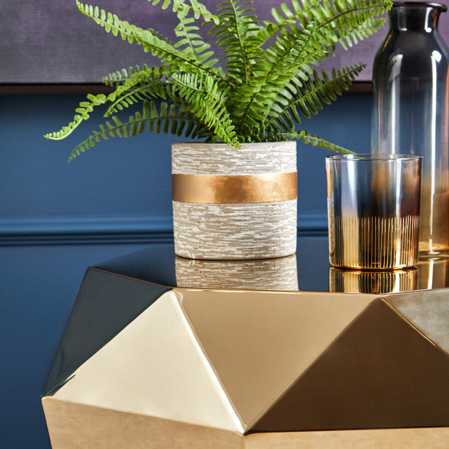 Geometric gold side table with a potted plant and glass on a blue wall background