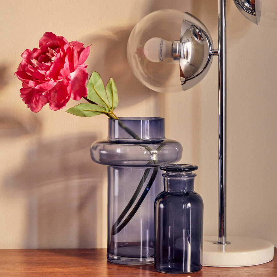 Glass vase with a pink flower on a wooden surface next to a lamp.