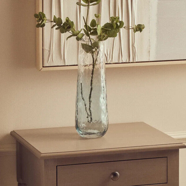 Wooden nightstand with a vase and books next to a bed with a pillow and magazine.