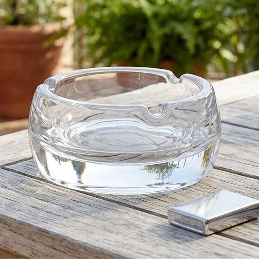 A clear glass smoking bowl sits on a weathered wooden table outdoors, with a silver lighter beside it and blurred potted plants in the background.