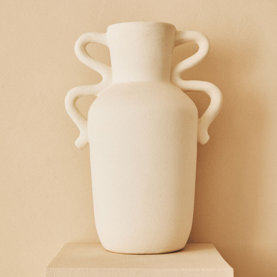 White vase with handles on a beige pedestal against a beige wall