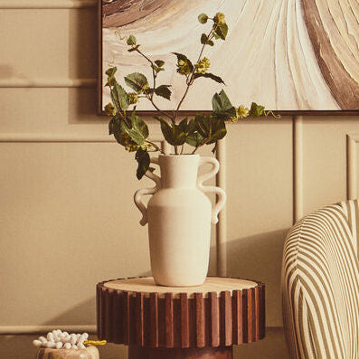 A framed abstract wall art print with swirling brown, beige, and cream marble-like patterns, displayed in a styled living room above a cream ribbed armchair and fluted wooden side tables.