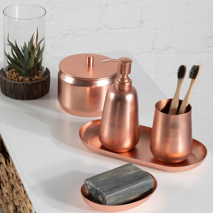 Copper bathroom accessories including a soap dish, bottle, and cup on a white surface with a plant in the background.