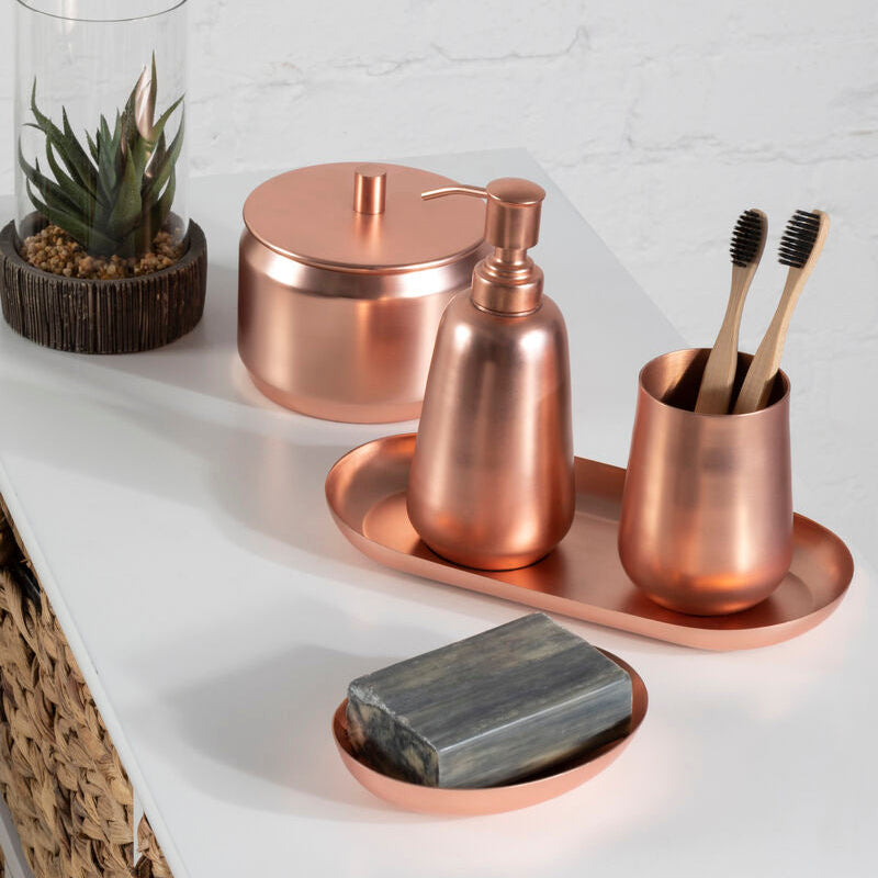 Copper bathroom accessories including a soap dish, bottle, and cup on a white surface with a plant in the background.