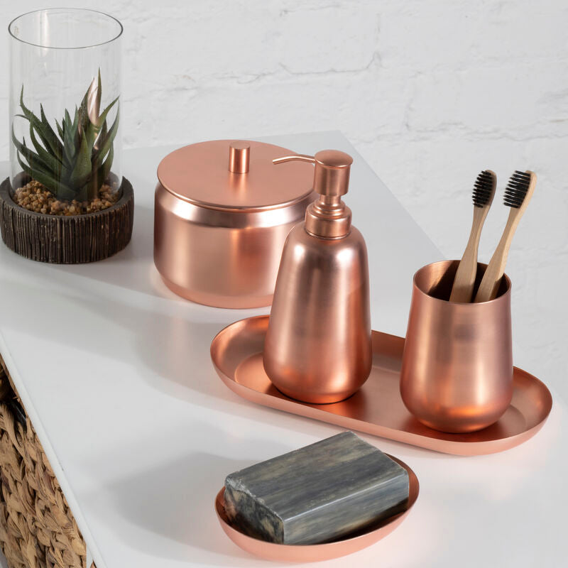 Copper bathroom accessories including a soap dish, bottle, and cup on a white surface with a plant in the background.
