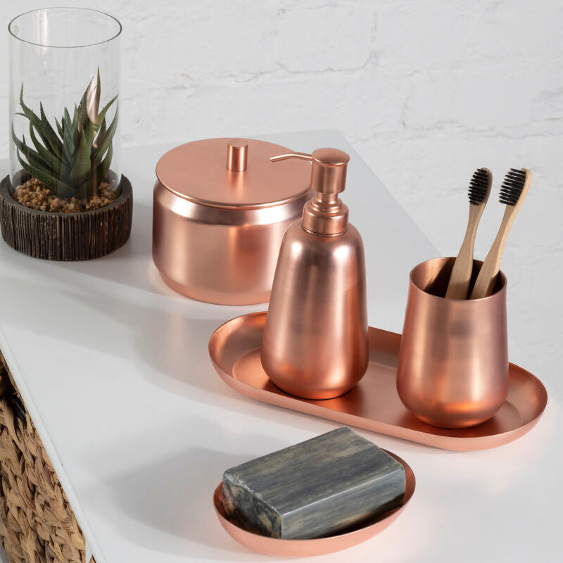 Copper bathroom accessories including a soap dish, bottle, and cup on a white surface with a plant in the background.