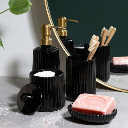 Black bathroom accessories including bottles, a soap dish, and toothbrush holder on a gray surface with a plant in the background.