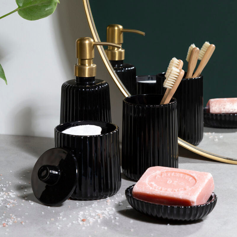 Black bathroom accessories including bottles, a soap dish, and toothbrush holder on a gray surface with a plant in the background.