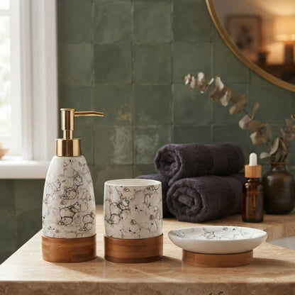 Three bathroom accessories in a marble-look finish with gold and wood accents, displayed on a countertop in a styled bathroom setting. The set includes a pump bottle, a tumbler, and a small dish, with folded towels, a bottle, and a mirror in the background.