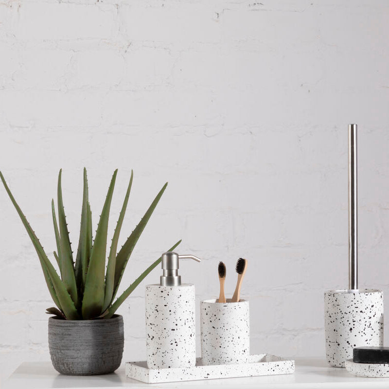 Bathroom accessories including a plant, soap dispenser, toothbrush holder, and toilet brush holder on a white surface with a light gray wall background.