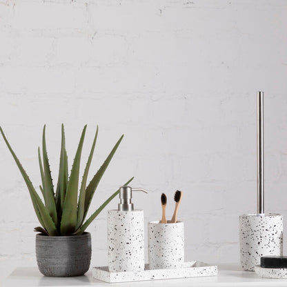 Bathroom accessories including a plant, soap dispenser, toothbrush holder, and toilet brush holder on a white surface with a light gray wall background.