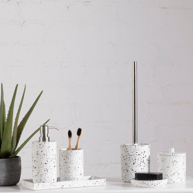 Bathroom accessories including a plant, soap dispenser, toothbrush holder, and toilet brush holder on a white surface with a light gray wall background.