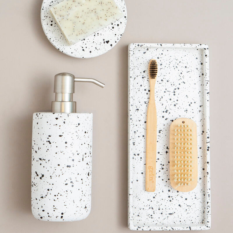 Speckled soap dispenser and tray with bamboo toothbrush and scrubber on a beige background