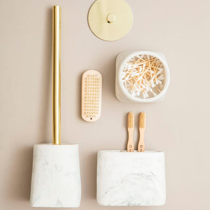 Bathroom accessories including a marble holder with a gold-plated brush, a small round holder with a candle, a white container with wooden toothbrushes, and a wooden scrubber on a beige background.
