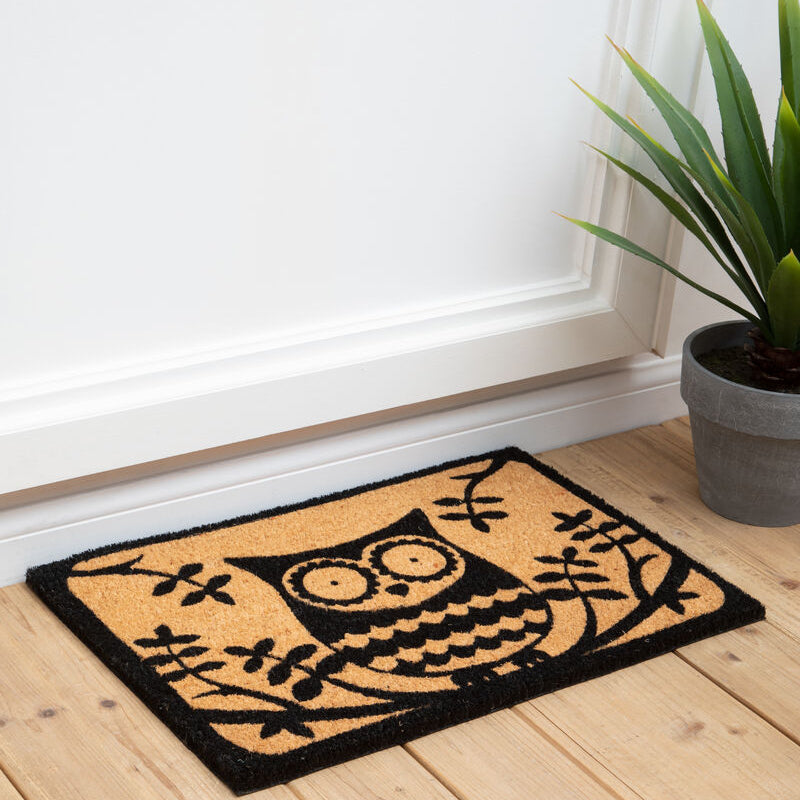 Doormat with owl design on a wooden floor next to a plant