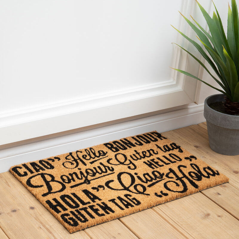 Doormat with multiple greetings on a wooden floor next to a plant