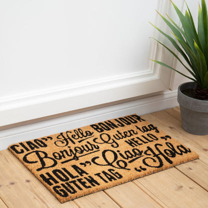 Doormat with multiple greetings on a wooden floor next to a plant
