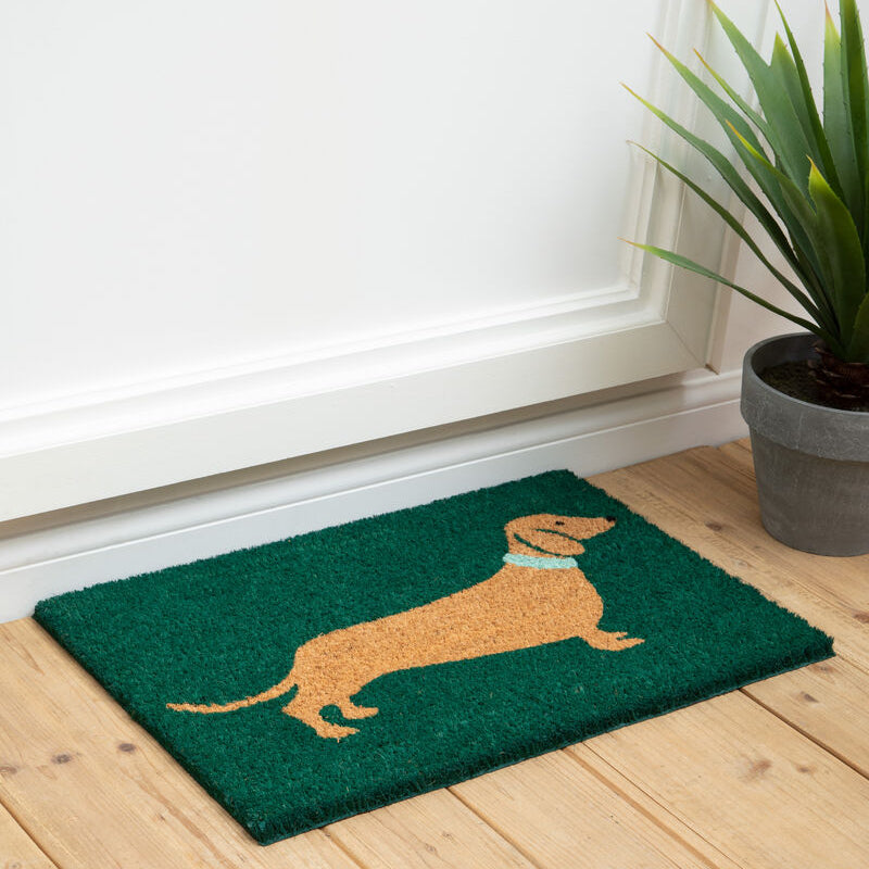 Green doormat with a brown dachshund design on a wooden floor.