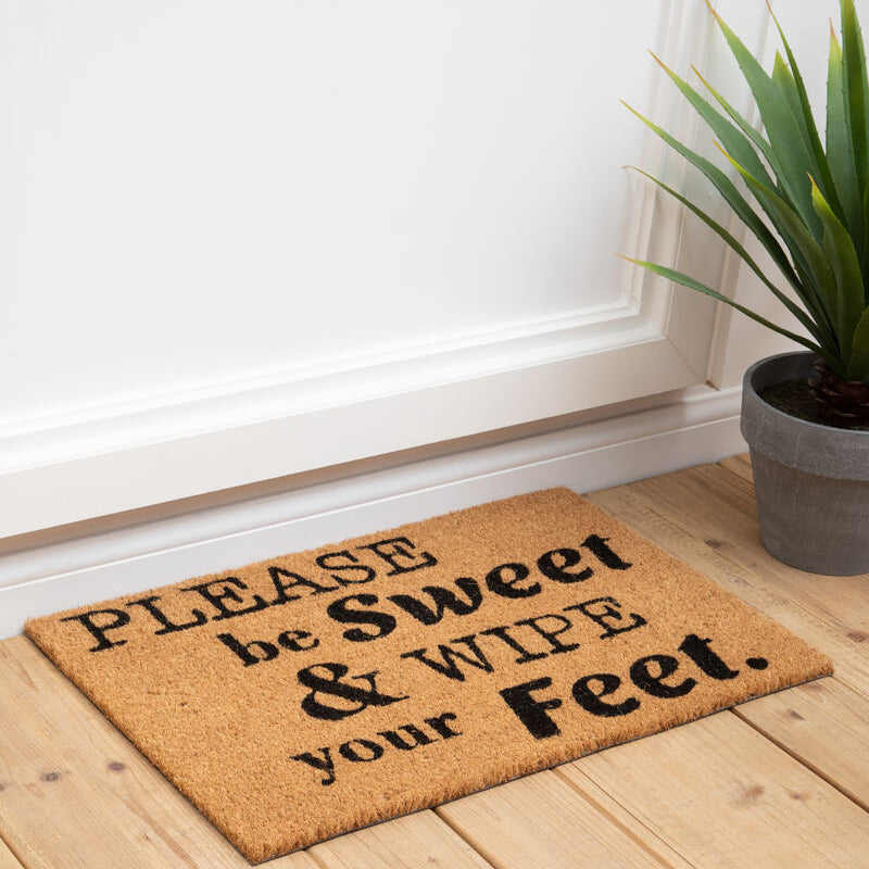 Doormat with humorous text on a wooden floor next to a plant