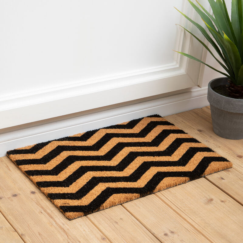 Doormat with zigzag pattern on a wooden floor next to a plant