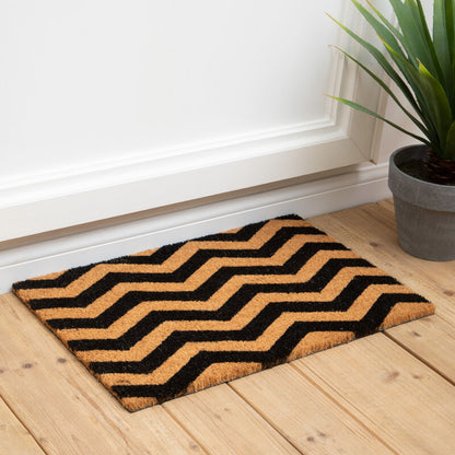 Doormat with zigzag pattern on a wooden floor next to a plant