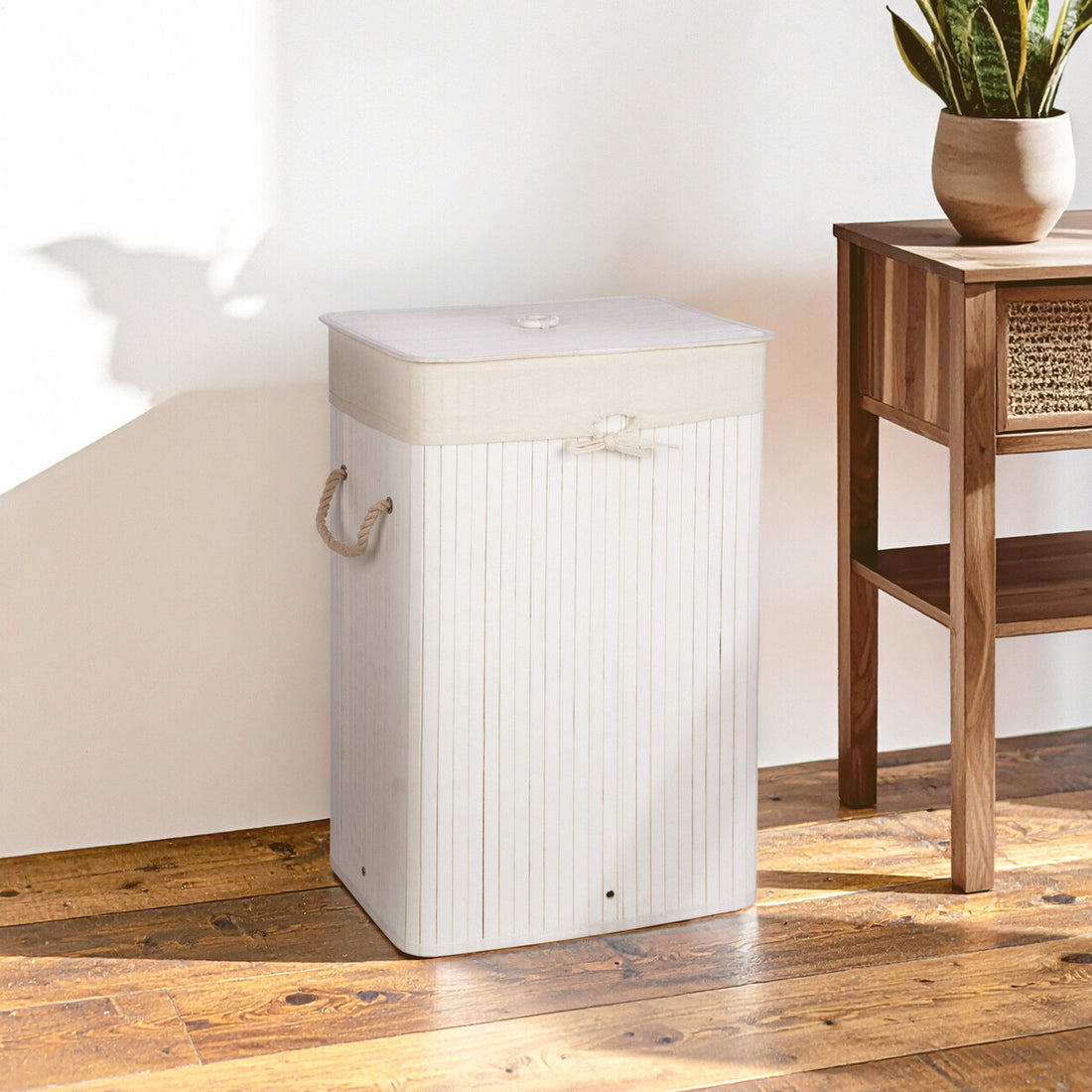 White laundry basket with a wooden handle on a wooden floor next to a wooden side table with a plant.