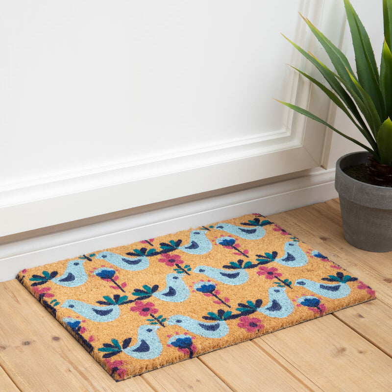 Decorative doormat with colorful pattern on a wooden floor next to a plant.