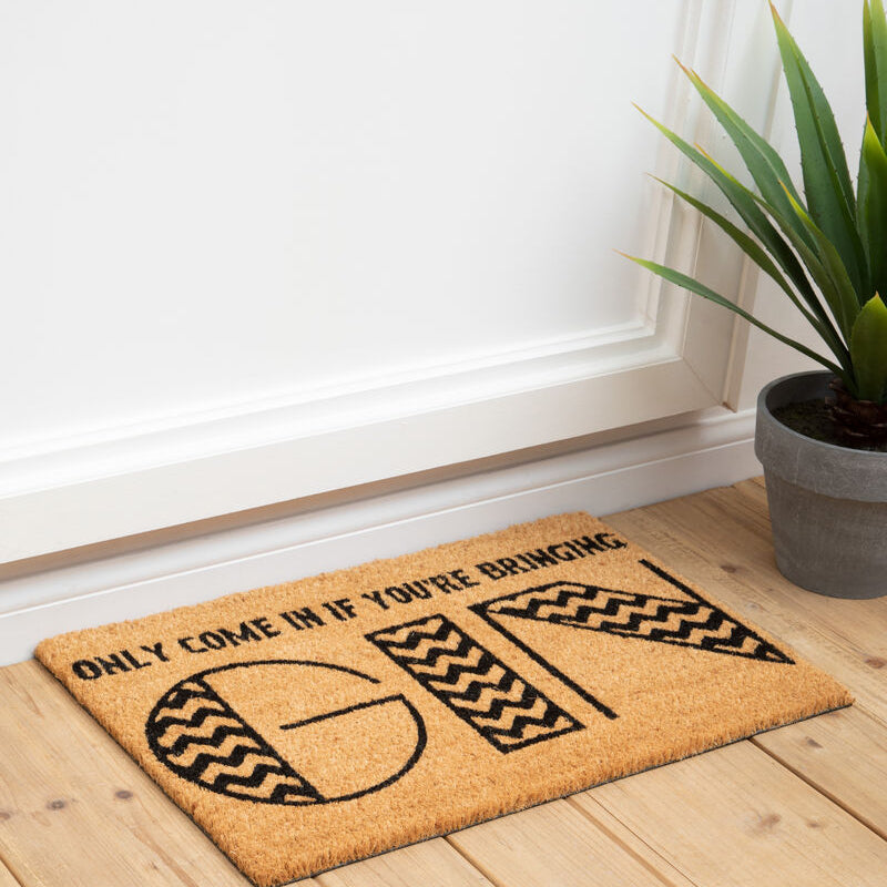 Doormat with humorous text on a wooden floor next to a plant