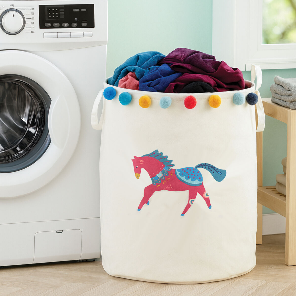 Laundry basket with colorful pom-poms and a horse design in front of a washing machine.