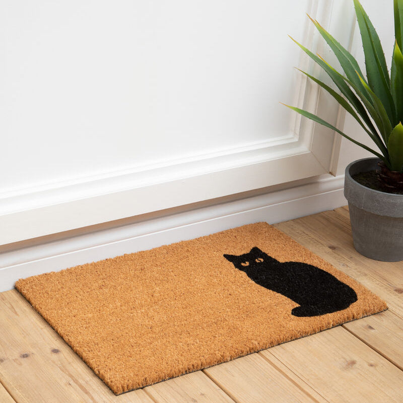 Doormat with a black cat design on a wooden floor next to a plant.